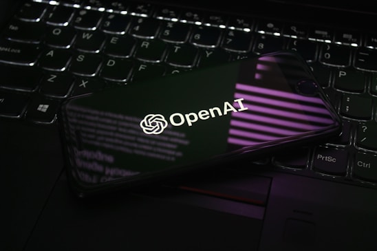 A smartphone displaying the OpenAI logo is resting on a laptop keyboard. The phone screen reflects purple and white light patterns, adding a modern and tech-focused ambiance.