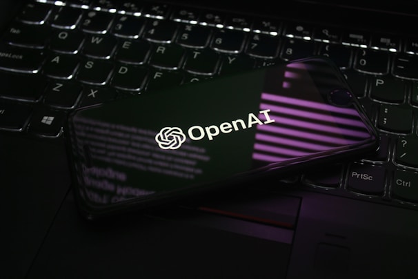 A smartphone displaying the OpenAI logo is resting on a laptop keyboard. The phone screen reflects purple and white light patterns, adding a modern and tech-focused ambiance.