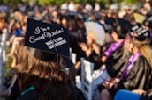 A group of people wearing black graduation caps and gowns sit outdoors, attending a graduation ceremony. The focus is on a cap with the phrase 'I'm a Social Worker! What's your superpower?' written on it. Sunlight casts a warm glow over the scene, highlighting the joyous atmosphere.