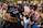 A group of people wearing black graduation caps and gowns sit outdoors, attending a graduation ceremony. The focus is on a cap with the phrase 'I'm a Social Worker! What's your superpower?' written on it. Sunlight casts a warm glow over the scene, highlighting the joyous atmosphere.