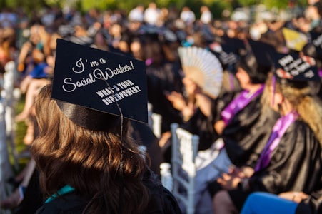 A group of people wearing black graduation caps and gowns sit outdoors, attending a graduation ceremony. The focus is on a cap with the phrase 'I'm a Social Worker! What's your superpower?' written on it. Sunlight casts a warm glow over the scene, highlighting the joyous atmosphere.