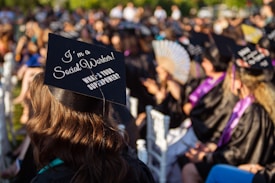 A group of people wearing black graduation caps and gowns sit outdoors, attending a graduation ceremony. The focus is on a cap with the phrase 'I'm a Social Worker! What's your superpower?' written on it. Sunlight casts a warm glow over the scene, highlighting the joyous atmosphere.