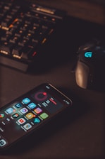 A smartphone displaying various social media app icons is placed on a dark surface. Nearby, a gaming mouse with a glowing logo and a mechanical keyboard with illuminated keys are visible. The lighting is low and focused, creating a moody atmosphere.