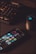 A smartphone displaying various social media app icons is placed on a dark surface. Nearby, a gaming mouse with a glowing logo and a mechanical keyboard with illuminated keys are visible. The lighting is low and focused, creating a moody atmosphere.