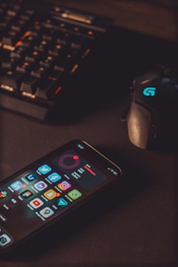 A smartphone displaying various social media app icons is placed on a dark surface. Nearby, a gaming mouse with a glowing logo and a mechanical keyboard with illuminated keys are visible. The lighting is low and focused, creating a moody atmosphere.