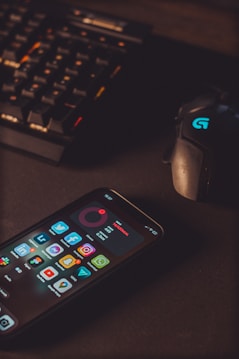 A smartphone displaying various social media app icons is placed on a dark surface. Nearby, a gaming mouse with a glowing logo and a mechanical keyboard with illuminated keys are visible. The lighting is low and focused, creating a moody atmosphere.