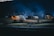 A group of tents with glowing lights is set up in a dark outdoor area, surrounded by bare trees under a deep blue night sky. The site looks like a camping area with a calm and peaceful atmosphere.