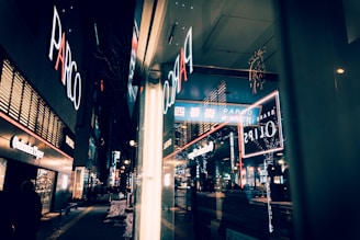 Night scene of an urban shopping district featuring bright neon signs and advertisements. The reflection in the glass creates a layered effect, adding complexity to the visual composition. The street is lined with illuminated shops, and pedestrians are visible in the background.