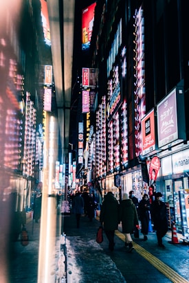 A bustling urban street scene at night with bright neon lights from signs illuminating the area. Crowds of people walk down the street, which is lined with shops and tall buildings. Reflections from the lights create a vibrant and colorful atmosphere.