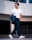 A fashionable man walks confidently, wearing sunglasses, a white t-shirt, dark trousers, and white sneakers. He holds a dark jacket draped over his forearm, and a silver chain necklace enhances his stylish appearance. The background features a modern, urban setting with architectural elements.