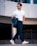 A fashionable man walks confidently, wearing sunglasses, a white t-shirt, dark trousers, and white sneakers. He holds a dark jacket draped over his forearm, and a silver chain necklace enhances his stylish appearance. The background features a modern, urban setting with architectural elements.