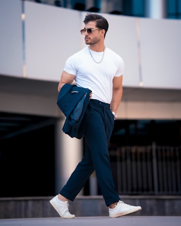 A fashionable man walks confidently, wearing sunglasses, a white t-shirt, dark trousers, and white sneakers. He holds a dark jacket draped over his forearm, and a silver chain necklace enhances his stylish appearance. The background features a modern, urban setting with architectural elements.