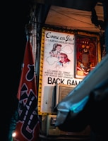 An illuminated retro-style poster hangs on a wall, depicting a man's and a woman's faces drawn in a vintage cartoon style. The poster bears the text 'Come on in! It's a Dream American Dream' and references a game of backgammon. A microphone is visible in front of the poster, and a red flag with partial lettering is in the foreground.