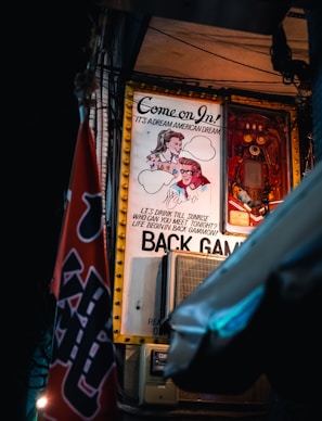 An illuminated retro-style poster hangs on a wall, depicting a man's and a woman's faces drawn in a vintage cartoon style. The poster bears the text 'Come on in! It's a Dream American Dream' and references a game of backgammon. A microphone is visible in front of the poster, and a red flag with partial lettering is in the foreground.