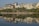 Modern buildings line the shoreline, reflected in the calm water of a lake. The image features mid-rise structures with glass facades and some greenery around them, highlighting urban development near the water's edge.
