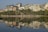 Modern buildings line the shoreline, reflected in the calm water of a lake. The image features mid-rise structures with glass facades and some greenery around them, highlighting urban development near the water's edge.