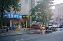 Two workers in orange uniforms are cleaning the street using hoses. They are near a row of parked cars including a red SUV. Behind them are storefronts with signage in Chinese characters and a large tree providing shade.