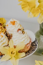 A decorative plate holding cupcakes topped with whipped cream and small decorative balls. Surrounding the cupcakes are vibrant yellow daffodils, adding a fresh and cheerful touch.