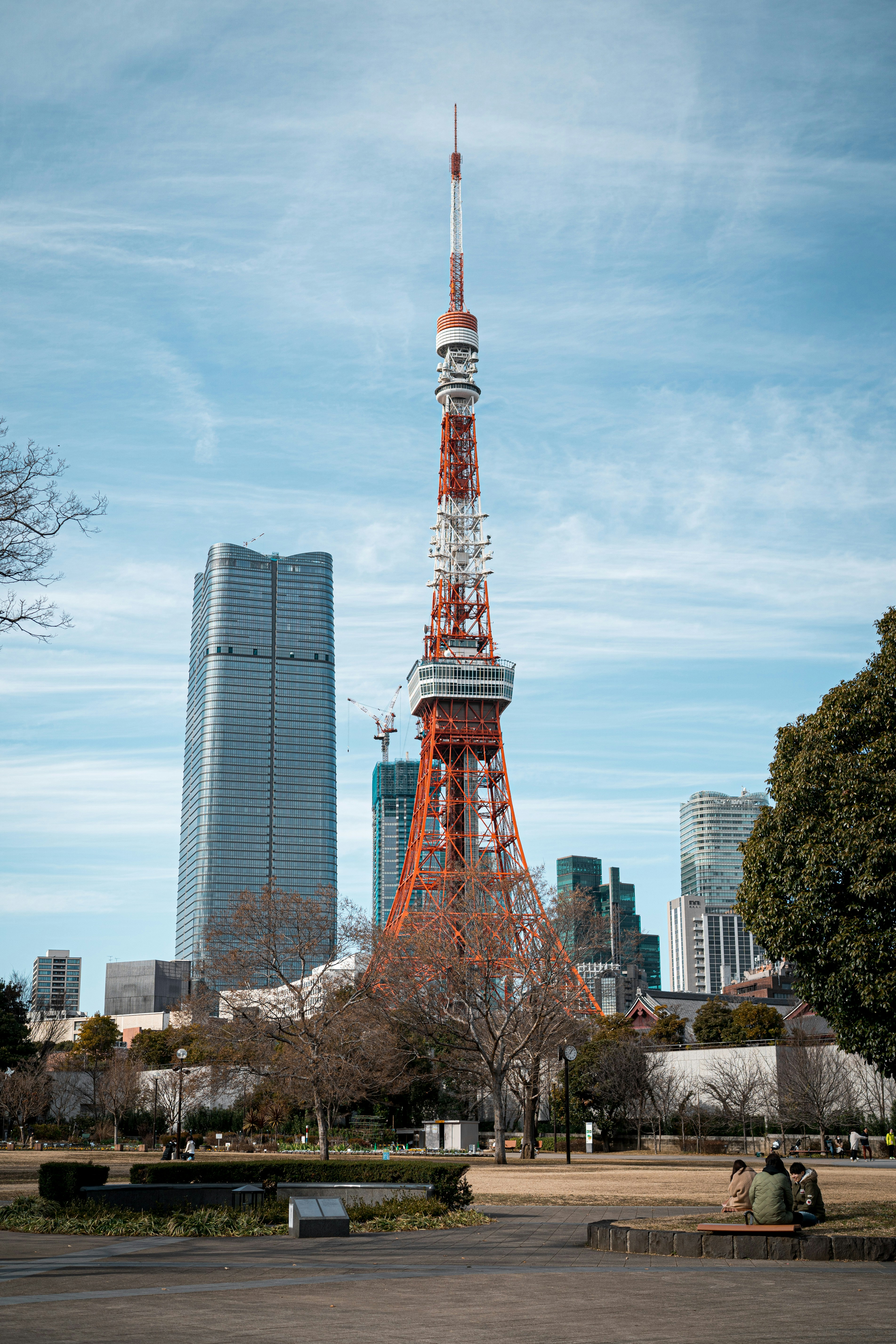 A large red and white broadcast tower stands prominently in an urban setting, surrounded by modern skyscrapers. Trees and a park are visible in the foreground, with a few people sitting and relaxing in the area.