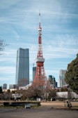 A large red and white broadcast tower stands prominently in an urban setting, surrounded by modern skyscrapers. Trees and a park are visible in the foreground, with a few people sitting and relaxing in the area.