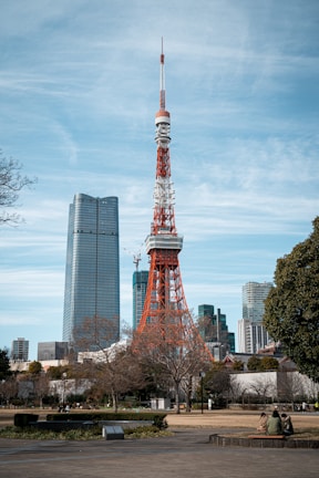 A large red and white broadcast tower stands prominently in an urban setting, surrounded by modern skyscrapers. Trees and a park are visible in the foreground, with a few people sitting and relaxing in the area.