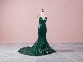 A dark green, sequined evening gown displayed on a mannequin. The gown features a form-fitting, mermaid silhouette with a dramatic train, set against a soft pink background and gray carpeted floor.