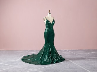 A dark green, sequined evening gown displayed on a mannequin. The gown features a form-fitting, mermaid silhouette with a dramatic train, set against a soft pink background and gray carpeted floor.