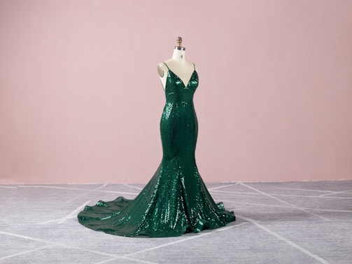 A dark green, sequined evening gown displayed on a mannequin. The gown features a form-fitting, mermaid silhouette with a dramatic train, set against a soft pink background and gray carpeted floor.