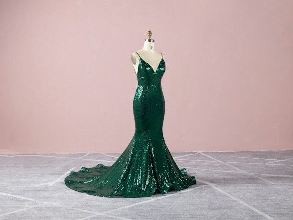 A dark green, sequined evening gown displayed on a mannequin. The gown features a form-fitting, mermaid silhouette with a dramatic train, set against a soft pink background and gray carpeted floor.