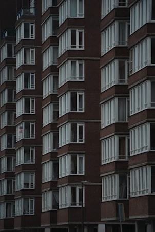 A series of modern, multi-story buildings with uniform brown brick facades and rows of identical white-framed windows. The architecture is clean and symmetrical, giving an impression of urban residential housing. The buildings are closely packed, creating a sense of density and order.