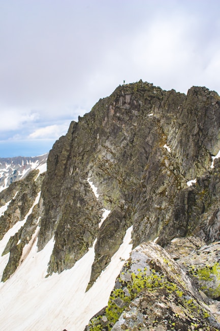 A rugged mountain peak with steep, rocky cliffs and patches of snow. One person is standing on top of the peak, surrounded by a vast, cloudy sky. The scene conveys a sense of adventure and solitude in a remote, high-altitude environment.