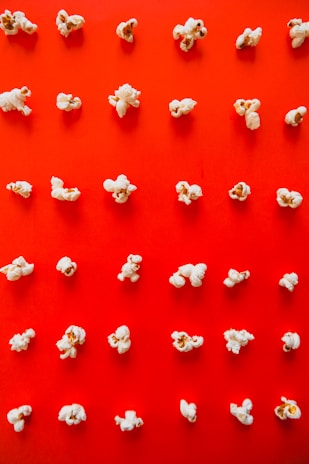 Popcorn is neatly arranged in a grid pattern on a bright red background, creating a visually striking contrast. Each kernel is distinct, with different shapes and sizes, highlighting the irregular nature of popcorn.