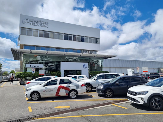 A modern commercial building with a prominent 'Consórcio Servopa' sign is surrounded by several parked cars. The glass facade of the dealership displays the Volkswagen logo. A partly cloudy sky and a casual outdoor setting create a busy yet calm environment.