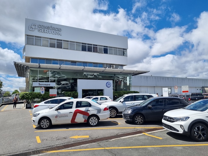 A modern commercial building with a prominent 'Consórcio Servopa' sign is surrounded by several parked cars. The glass facade of the dealership displays the Volkswagen logo. A partly cloudy sky and a casual outdoor setting create a busy yet calm environment.