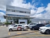 A modern commercial building with a prominent 'Cons&oacute;rcio Servopa' sign is surrounded by several parked cars. The glass facade of the dealership displays the Volkswagen logo. A partly cloudy sky and a casual outdoor setting create a busy yet calm environment.