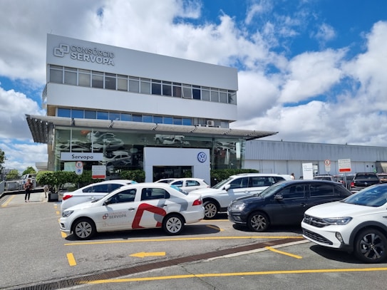 A modern commercial building with a prominent 'Cons&oacute;rcio Servopa' sign is surrounded by several parked cars. The glass facade of the dealership displays the Volkswagen logo. A partly cloudy sky and a casual outdoor setting create a busy yet calm environment.
