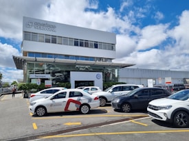 A modern commercial building with a prominent 'Consórcio Servopa' sign is surrounded by several parked cars. The glass facade of the dealership displays the Volkswagen logo. A partly cloudy sky and a casual outdoor setting create a busy yet calm environment.