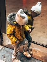 A child wearing a yellow jacket with a fur-lined hood and a white knit hat is looking down while standing in front of a reflective surface, possibly a glass door. The reflection shows the child’s mirror image. The child is also wearing light pants and sneakers with Velcro straps.