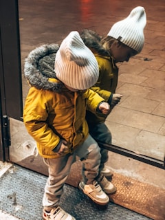 A child wearing a yellow jacket with a fur-lined hood and a white knit hat is looking down while standing in front of a reflective surface, possibly a glass door. The reflection shows the child’s mirror image. The child is also wearing light pants and sneakers with Velcro straps.
