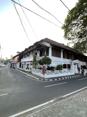 A large white colonial-style building with dark wooden shutters and a tiled roof sits at the corner of an urban street. The building is surrounded by lush green plants and a white fence. The street is wide and clear, with overhead power lines crisscrossing above. Trees with thick foliage provide some shade.