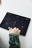 A person is playing a retro-style space video game on a tablet. The player's finger is seen interacting with the on-screen controls, which include direction arrows and possibly a fire button. The game appears to involve controlling a spaceship avoiding or attacking obstacles like asteroids or enemy ships.