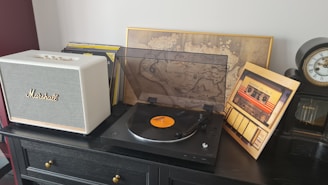 A stylish home setup featuring a white Marshall speaker on a wooden table, accompanied by a modern turntable with a vinyl record. The table also holds a vintage-style map framed in gold and a classic analog clock. A display of vinyl album covers adds a retro aesthetic.
