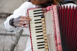 A person is playing a Hohner accordion, with fingers visibly pressing on the keys. The instrument has a red and cream color scheme, with intricate details and patterns on its body. The person has a tattooed hand adorned with rings, and a watch is visible on the wrist. The background is blurred, focusing attention on the accordion and the hand.