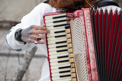 A person is playing a Hohner accordion, with fingers visibly pressing on the keys. The instrument has a red and cream color scheme, with intricate details and patterns on its body. The person has a tattooed hand adorned with rings, and a watch is visible on the wrist. The background is blurred, focusing attention on the accordion and the hand.