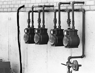 A black and white image of four large, industrial gas meters mounted on a concrete wall with visible piping and connections. The setup is located indoors or in an industrial area, and a section of brickwork is visible above the concrete.