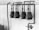 A black and white image of four large, industrial gas meters mounted on a concrete wall with visible piping and connections. The setup is located indoors or in an industrial area, and a section of brickwork is visible above the concrete.