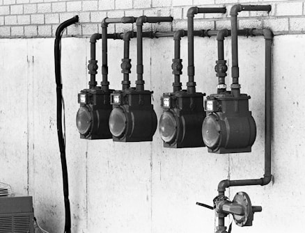 A black and white image of four large, industrial gas meters mounted on a concrete wall with visible piping and connections. The setup is located indoors or in an industrial area, and a section of brickwork is visible above the concrete.