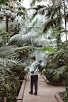 A person standing in a lush greenhouse, surrounded by various large green plants and palm trees. The setting is serene with natural light filtering through the glass structure. The person is wearing casual clothing and carrying a backpack, gazing at the foliage.