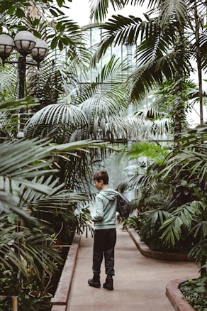 A person standing in a lush greenhouse, surrounded by various large green plants and palm trees. The setting is serene with natural light filtering through the glass structure. The person is wearing casual clothing and carrying a backpack, gazing at the foliage.