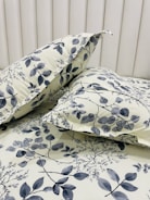 Two pillows are placed on a bed with a matching floral-patterned fabric. The fabric features a botanical design with leaves and branches in shades of gray against a light cream background. The headboard is tufted and covered in a soft, light-colored material.